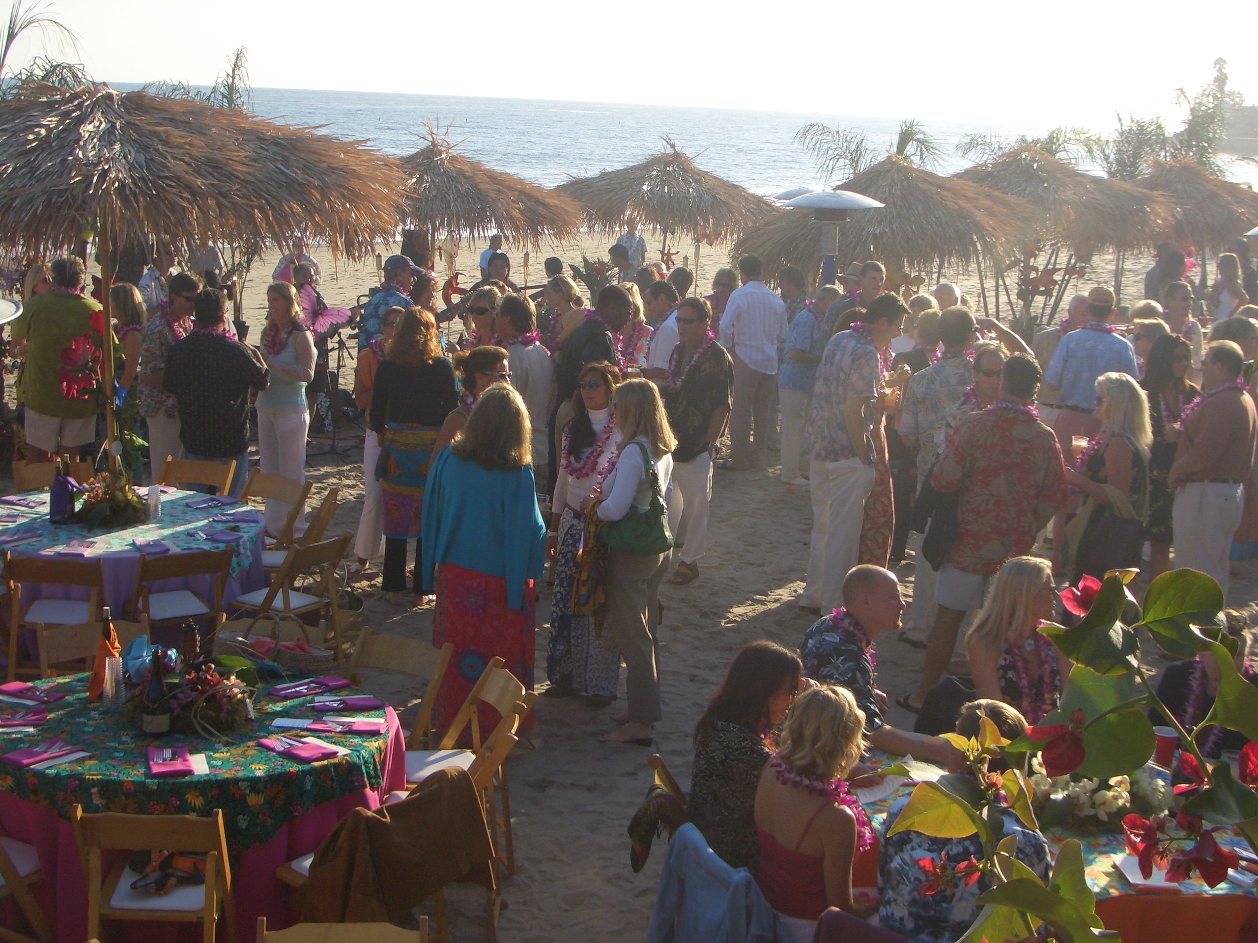Large beach luau event with thatched umbrellas, colorful tablecloths, and crowd at sunset in San Diego, CA